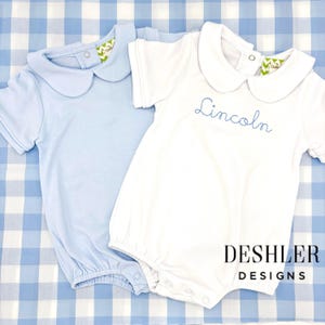 May include: Two baby rompers, one light blue and one white, with Peter Pan collars. The white romper has the name "Lincoln" embroidered in blue. The rompers are on a blue and white checkered background. The brand name "DESHLER DESIGNS" is printed in black.