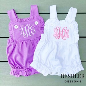 May include: Two baby rompers, one lavender and one white, with ruffled edges and button details. Each romper features a monogram in a contrasting color. The lavender romper has a white monogram, and the white romper has a pink monogram. The bottom of each romper has snaps.