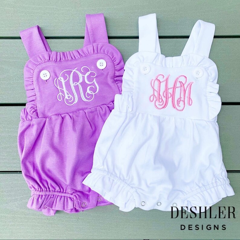 May include: Two baby rompers, one lavender and one white, with ruffled edges and button details. Each romper features a monogram in a contrasting color. The lavender romper has a white monogram, and the white romper has a pink monogram. The bottom of each romper has snaps.