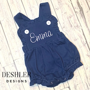 May include: Navy blue baby romper with white embroidered name "Emma". The romper features ruffled edges, white buttons, and snap closures. The Deshler Designs logo is visible at the bottom left.