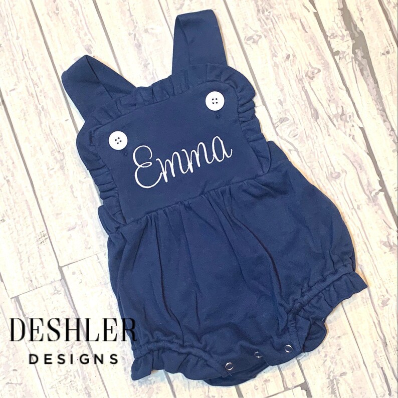 May include: Navy blue baby romper with white embroidered name "Emma". The romper features ruffled edges, white buttons, and snap closures. The Deshler Designs logo is visible at the bottom left.