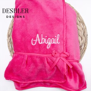 May include: A bright pink baby blanket with the name "Abigail" embroidered in white script. The blanket features a ruffled edge and a decorative bow. The blanket is from Deshler Designs.