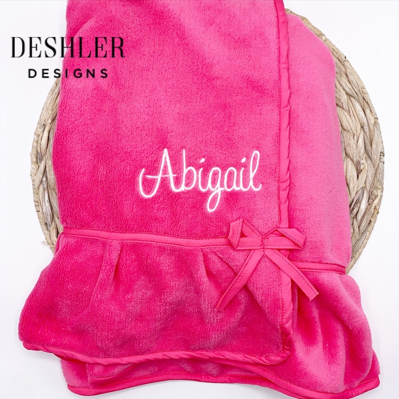 May include: A bright pink baby blanket with the name "Abigail" embroidered in white script. The blanket features a ruffled edge and a decorative bow. The blanket is from Deshler Designs.