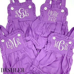 May include: Four purple baby rompers with white embroidered initials and names. Each romper has ruffled edges, white buttons, and adjustable straps. The text "DESHLER DESIGNS" is printed in black.