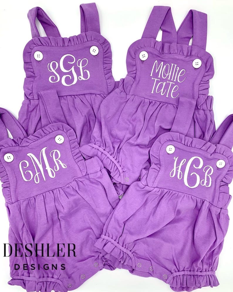 May include: Four purple baby rompers with white embroidered initials and names. Each romper has ruffled edges, white buttons, and adjustable straps. The text "DESHLER DESIGNS" is printed in black.