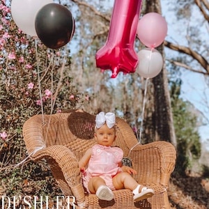 May include: A baby in a pink romper with a white bow headband sits in a wicker chair, surrounded by balloons. A large, pink number one balloon floats above. Other balloons are white and black. The image has the text "DESHLER DESIGNS" at the bottom.