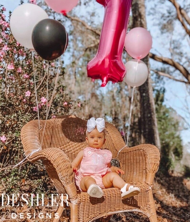 May include: A baby in a pink romper with a white bow headband sits in a wicker chair, surrounded by balloons. A large, pink number one balloon floats above. Other balloons are white and black. The image has the text "DESHLER DESIGNS" at the bottom.