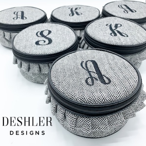 May include: Set of six round black and white patterned fabric storage boxes with black zipper closures and ruffled edges. Each box has a different letter monogrammed on the top in black thread. The boxes are arranged in a circle on a white surface. The text "DESHLER DESIGNS" is printed below the boxes.
