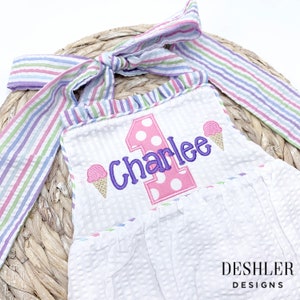 May include: A white onesie with a pink and white number one and the name "Charlee" embroidered in purple. The onesie has a pink and white striped ruffle around the neckline and a pink and white striped bow tie. The onesie is decorated with two pink and white ice cream cones.