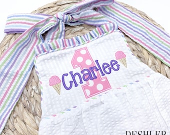 Puede incluir: Un pelele blanco con un número uno rosa y blanco y el nombre "Charlee" bordado en morado. El pelele tiene un volante rosa y blanco a rayas alrededor del cuello y una pajarita rosa y blanca a rayas. El pelele está decorado con dos conos de helado rosa y blanco.