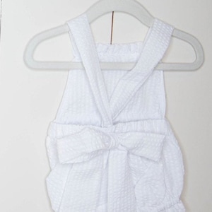 May include: White baby romper with a bow at the waist. The romper is made of a soft, textured fabric and has a sleeveless design.