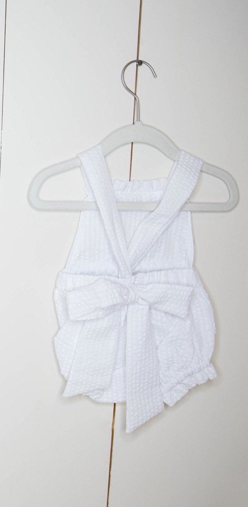 May include: White baby romper with a bow at the waist. The romper is made of a soft, textured fabric and has a sleeveless design.