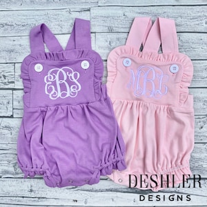 May include: Two baby rompers, one lavender and one pink, with white embroidered monograms. Each romper has ruffled trim, button details, and DESHLER DESIGNS printed at the bottom.
