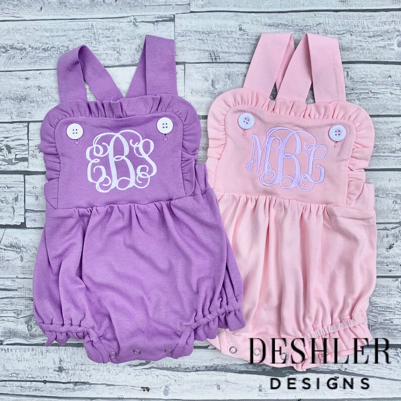 May include: Two baby rompers, one lavender and one pink, with white embroidered monograms. Each romper has ruffled trim, button details, and DESHLER DESIGNS printed at the bottom.