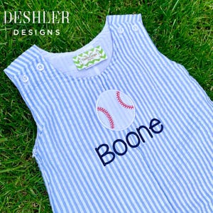 May include: A blue and white striped baby onesie with a baseball and the name "Boone" embroidered on the front.
