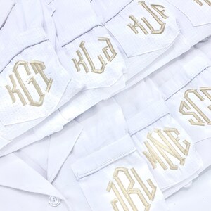 May include: White pajama shirts with embroidered initials in a gold color. The shirts are neatly stacked, showcasing the personalized monogram details on the pocket. The image also displays the "DESHLER DESIGNS" logo.