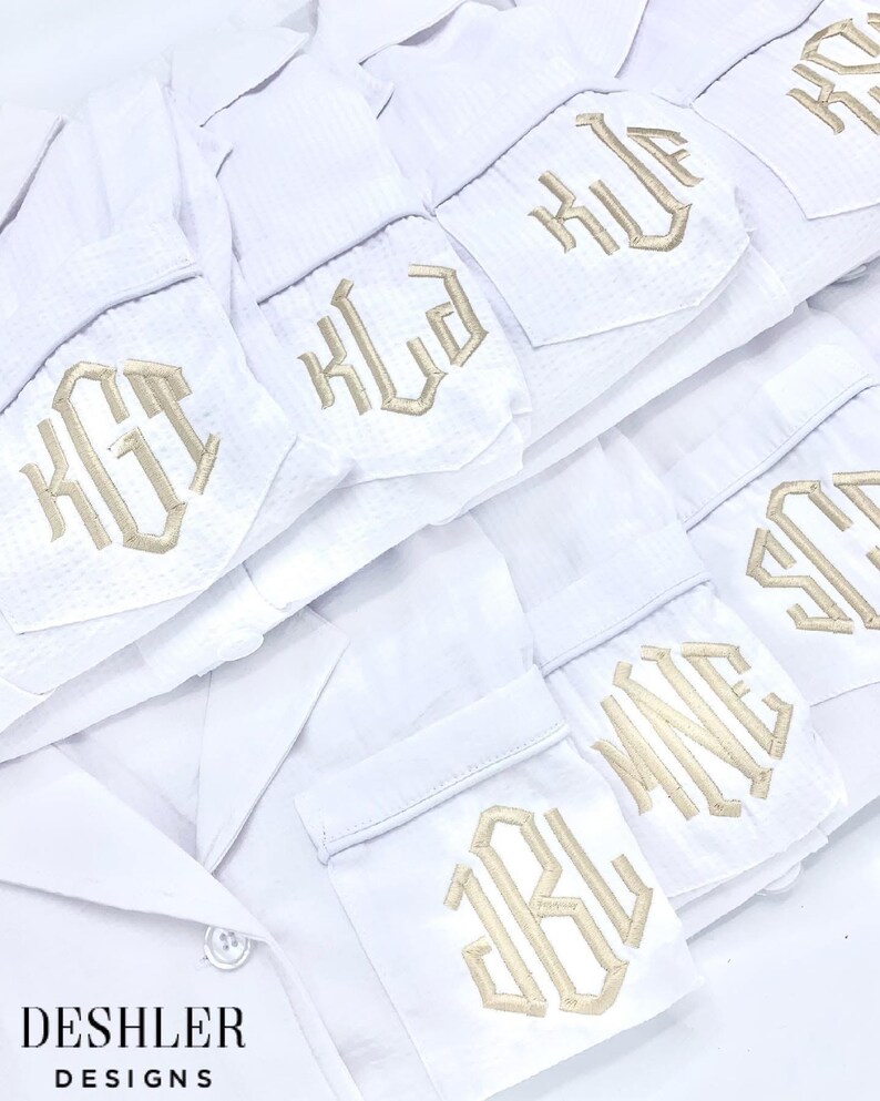 May include: White pajama shirts with embroidered initials in a gold color. The shirts are neatly stacked, showcasing the personalized monogram details on the pocket. The image also displays the "DESHLER DESIGNS" logo.