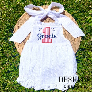 May include: White onesie with red, white and blue stripes, a number one, and the name Gracie embroidered on the front. The onesie has a ruffled bottom and long sleeves.