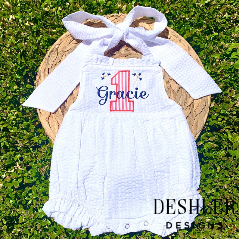 May include: White onesie with red, white and blue stripes, a number one, and the name Gracie embroidered on the front. The onesie has a ruffled bottom and long sleeves.