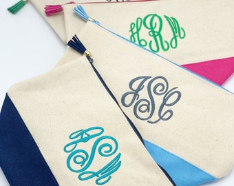 May include: Four canvas zippered pouches with different colored fabric accents and embroidered monograms. The pouches are arranged in a stack with the top pouch showing a green accent, the next pouch showing a pink accent, the next pouch showing a blue accent, and the bottom pouch showing a navy blue accent. The pouches are all white with a zipper closure and a tassel.