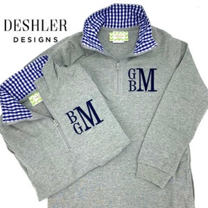 May include: Two gray quarter-zip pullovers with navy blue monogrammed initials. The collars and inner cuffs feature a blue and white gingham pattern. The brand name "Deshler Designs" is printed in black.
