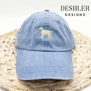 May include: A light blue denim baseball cap with a beige embroidered dog silhouette. The cap has a curved brim and a structured crown. The text "DESHLER DESIGNS" is printed in black on the back of the cap.