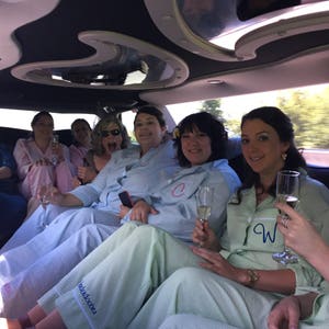 May include: A group of people in a limousine, wearing matching pajamas and holding champagne flutes. The pajamas are light blue and green striped, with some having initials embroidered on the chest. The interior of the limousine is dark with a mirrored ceiling.