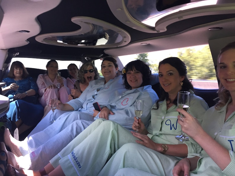 May include: A group of people in a limousine, wearing matching pajamas and holding champagne flutes. The pajamas are light blue and green striped, with some having initials embroidered on the chest. The interior of the limousine is dark with a mirrored ceiling.