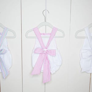 Op de afbeelding: Drie witte seersucker rompers met strikdetails hangen aan grijze hangers. E&eacute;n romper heeft een gestreepte strik met pastelkleuren, een andere heeft een roze gestreepte strik en de derde heeft een witte strik.