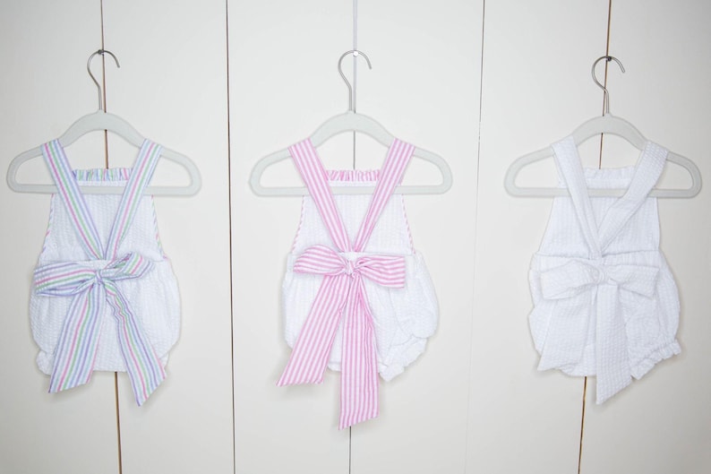 Op de afbeelding: Drie witte seersucker rompers met strikdetails hangen aan grijze hangers. E&eacute;n romper heeft een gestreepte strik met pastelkleuren, een andere heeft een roze gestreepte strik en de derde heeft een witte strik.