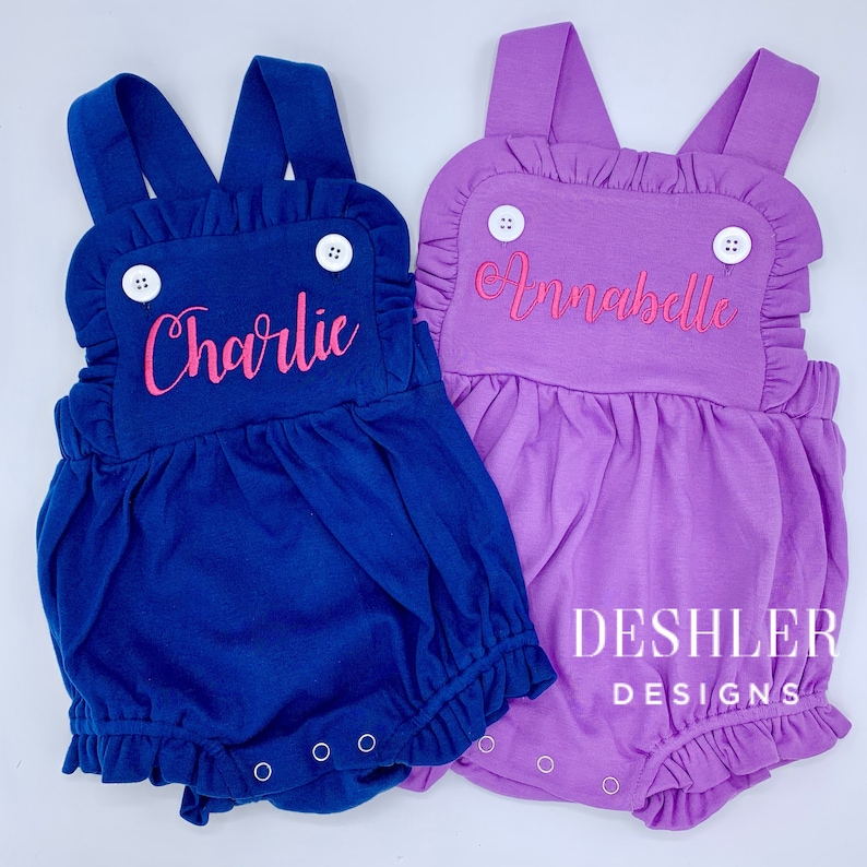 May include: Two baby rompers, one navy blue and one lavender, with ruffled details and suspender straps. Each romper has a name embroidered in pink: "Charlie" and "Annabelle". White buttons and snap closures are visible. The brand name "DESHLER DESIGNS" is printed on the bottom right.