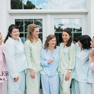 May include: A group of women wearing matching pastel-colored pajama sets, each with a monogrammed initial on the shirt. The pajamas are light pink, blue, and green. The image includes the text "DESHLER DESIGNS".