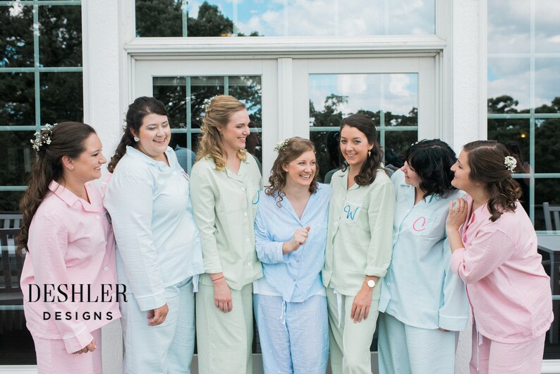 May include: A group of women wearing matching pastel-colored pajama sets, each with a monogrammed initial on the shirt. The pajamas are light pink, blue, and green. The image includes the text "DESHLER DESIGNS".