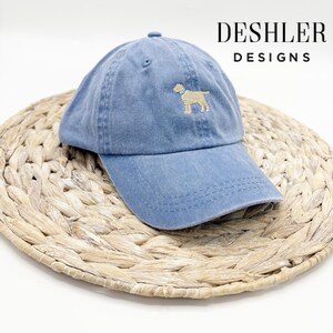 May include: A blue denim baseball cap with a beige embroidered dog design. The cap has a curved brim and an adjustable strap. The text "DESHLER DESIGNS" is visible in the upper right corner of the image.