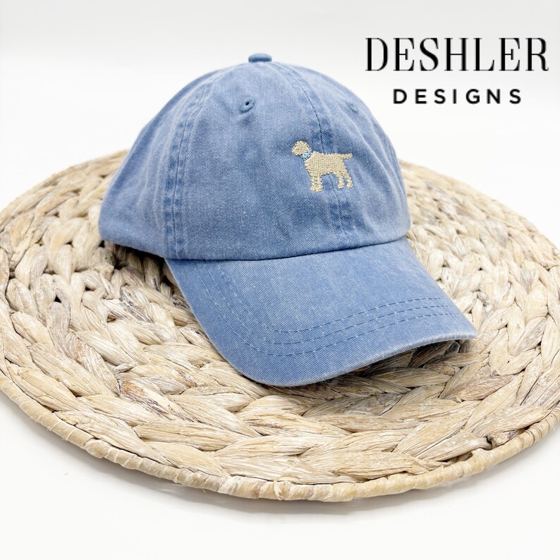 May include: A blue denim baseball cap with a beige embroidered dog design. The cap has a curved brim and an adjustable strap. The text "DESHLER DESIGNS" is visible in the upper right corner of the image.