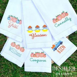 May include: Six white kitchen towels with embroidered designs. The designs include pumpkins, a truck, and a row of boats. Each towel has a name embroidered below the design. The names are Grisi, Gleason, Connelly, Williams, Cosgrove, and Bien.