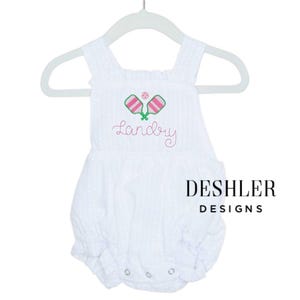 Op de afbeelding: Witte seersucker romper met geborduurde roze en groene pickleball rackets en een roze pickleball. De naam "Landry" is geborduurd in roze script. De tekst "DESHLER DESIGNS" is in zwart gedrukt.