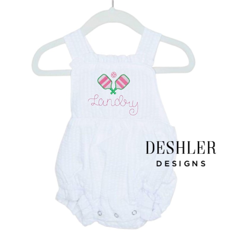 Op de afbeelding: Witte seersucker romper met geborduurde roze en groene pickleball rackets en een roze pickleball. De naam "Landry" is geborduurd in roze script. De tekst "DESHLER DESIGNS" is in zwart gedrukt.