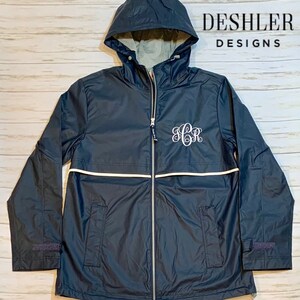 May include: A navy blue hooded rain jacket with a white stripe and a white monogrammed "SCR" on the front. The jacket has a zipper closure and two pockets. The text "DESHLER DESIGNS" is printed on the back of the jacket.