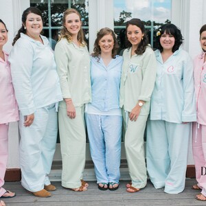 May include: Group of people wearing matching pajama sets in various pastel colors. The pajamas include button-down shirts and pants. Some shirts have embroidered initials. The image is taken outdoors.