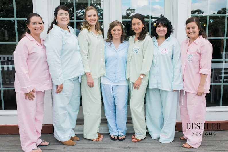 May include: Group of people wearing matching pajama sets in various pastel colors. The pajamas include button-down shirts and pants. Some shirts have embroidered initials. The image is taken outdoors.
