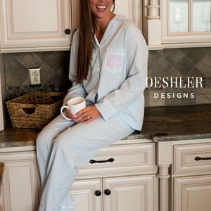 May include: Light blue and white striped pajama set with a monogram on the chest pocket. The pajama top has a button-down front and a collar. The matching pants are also striped. The text "DESHLER DESIGNS" is visible.