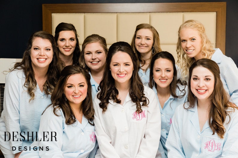 May include: A group of smiling people wearing light blue and white striped pajama shirts. One person wears a white pajama shirt with a pink monogram. The logo "DESHLER DESIGNS" is visible in the bottom left corner.