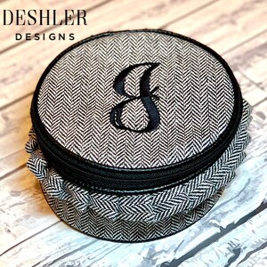 May include: A round, black and white,  herringbone patterned fabric jewelry box with a black zipper and a black embroidered letter "J" on the lid.