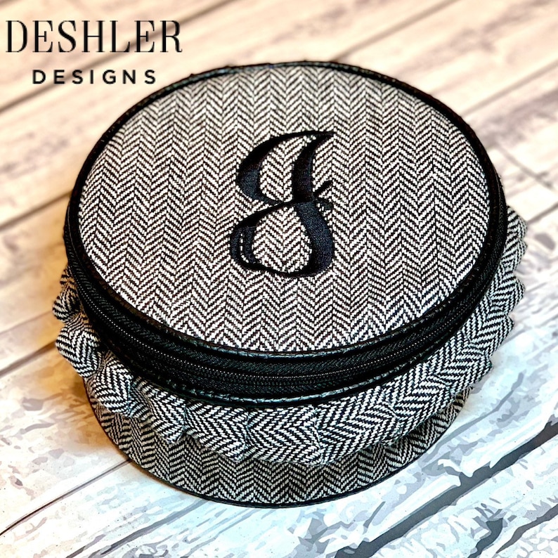 May include: A round, black and white,  herringbone patterned fabric jewelry box with a black zipper and a black embroidered letter "J" on the lid.