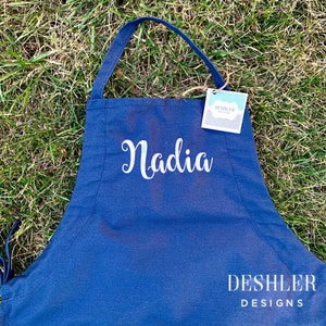 May include: A navy blue apron with the name "Nadia" embroidered in white on the front. The apron has adjustable straps and a pocket. The apron is made by Deshler Designs.