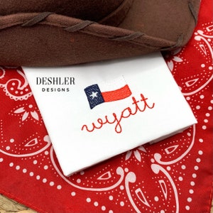 May include: White fabric with red embroidery of the name "wyatt" and a small embroidered Texas flag. The fabric is on top of a red bandana with white polka dots.
