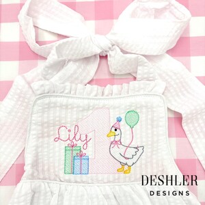 May include: White seersucker birthday dress with a large bow. The dress features an embroidered design with the name "Lily", the number "1", a goose with a party hat, and two gift boxes. The text "DESHLER DESIGNS" is at the bottom.