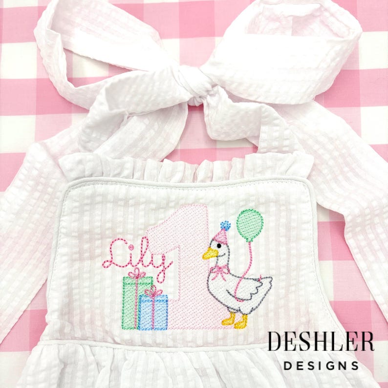 May include: White seersucker birthday dress with a large bow. The dress features an embroidered design with the name "Lily", the number "1", a goose with a party hat, and two gift boxes. The text "DESHLER DESIGNS" is at the bottom.