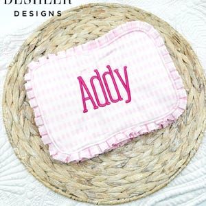 May include: A pink and white gingham baby blanket with the name "Addy" embroidered in a bright pink script. The blanket has a ruffled edge and is set on a woven placemat. The text "Deshler Designs" is in the upper left corner.